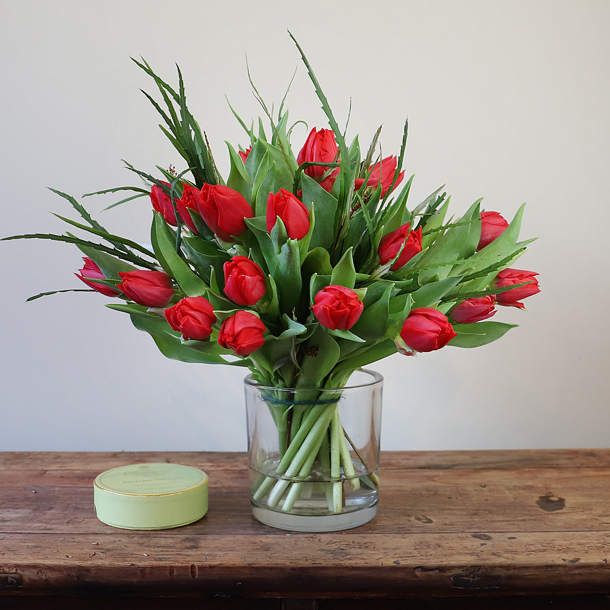 A bouquet of red tulips and foilage in a glass vase with a box of chocolates