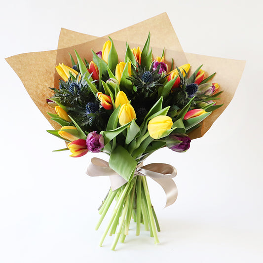 A bouqet of yellow, orange and dark purple tulips with blue thistle wrapped in brown paper