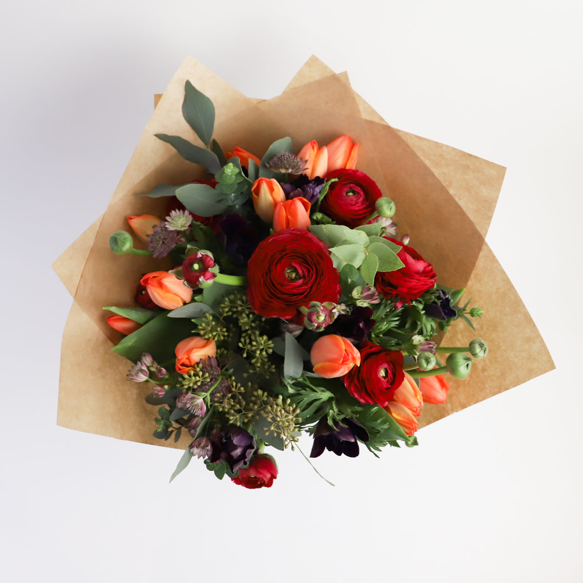 Overhead view of red ranunulus, blue thistle, orange and dark purple tulips with seasonal foliage