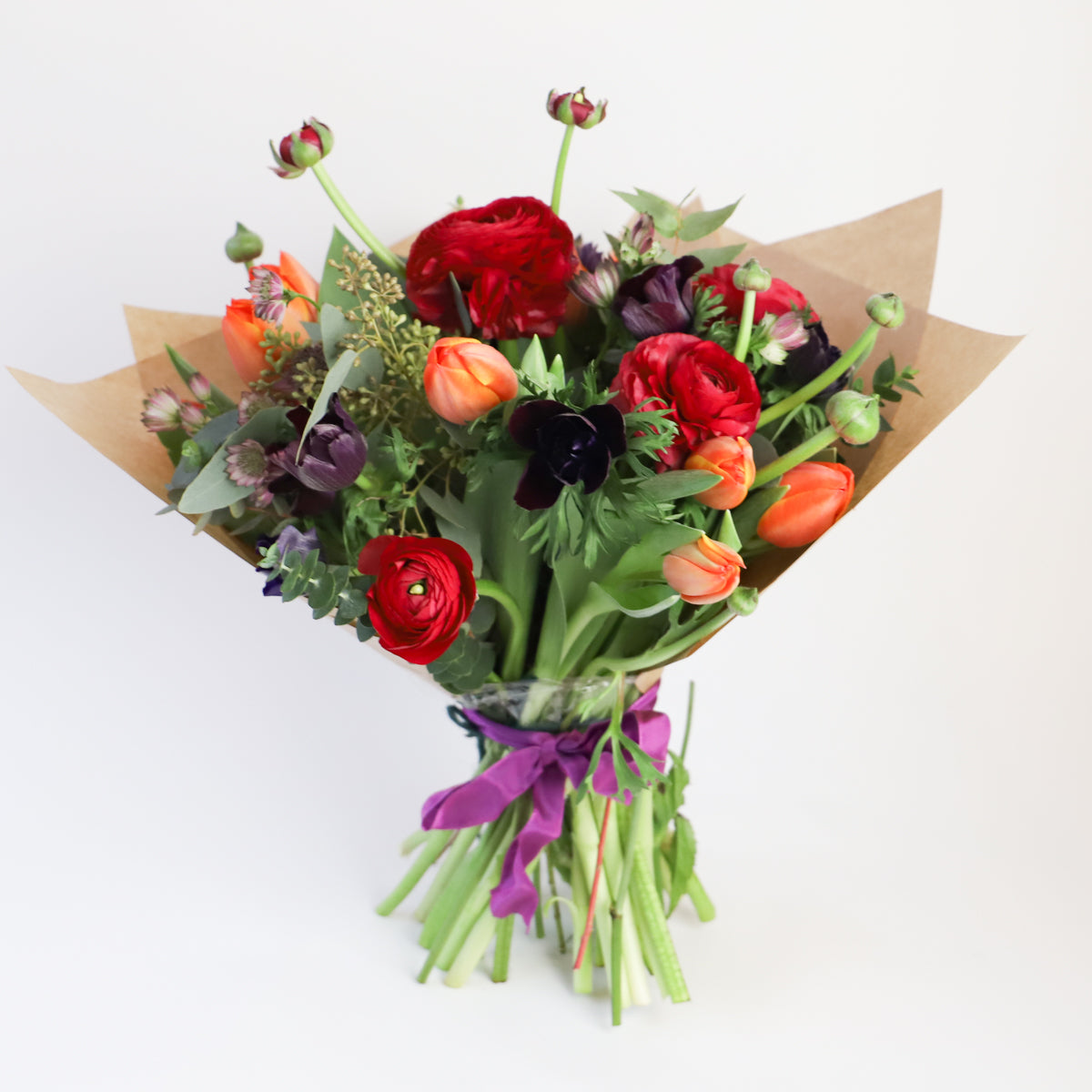 A bouquet containing red ranunulus, blue thistle, orange and dark purple tulips with seasonal foliage