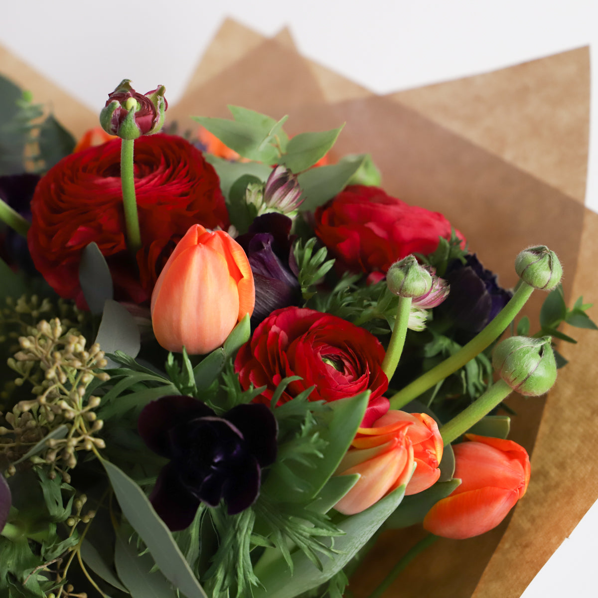 Detail close up of bouquet containing red ranunulus, blue thistle, orange and dark purple tulips with seasonal foliage