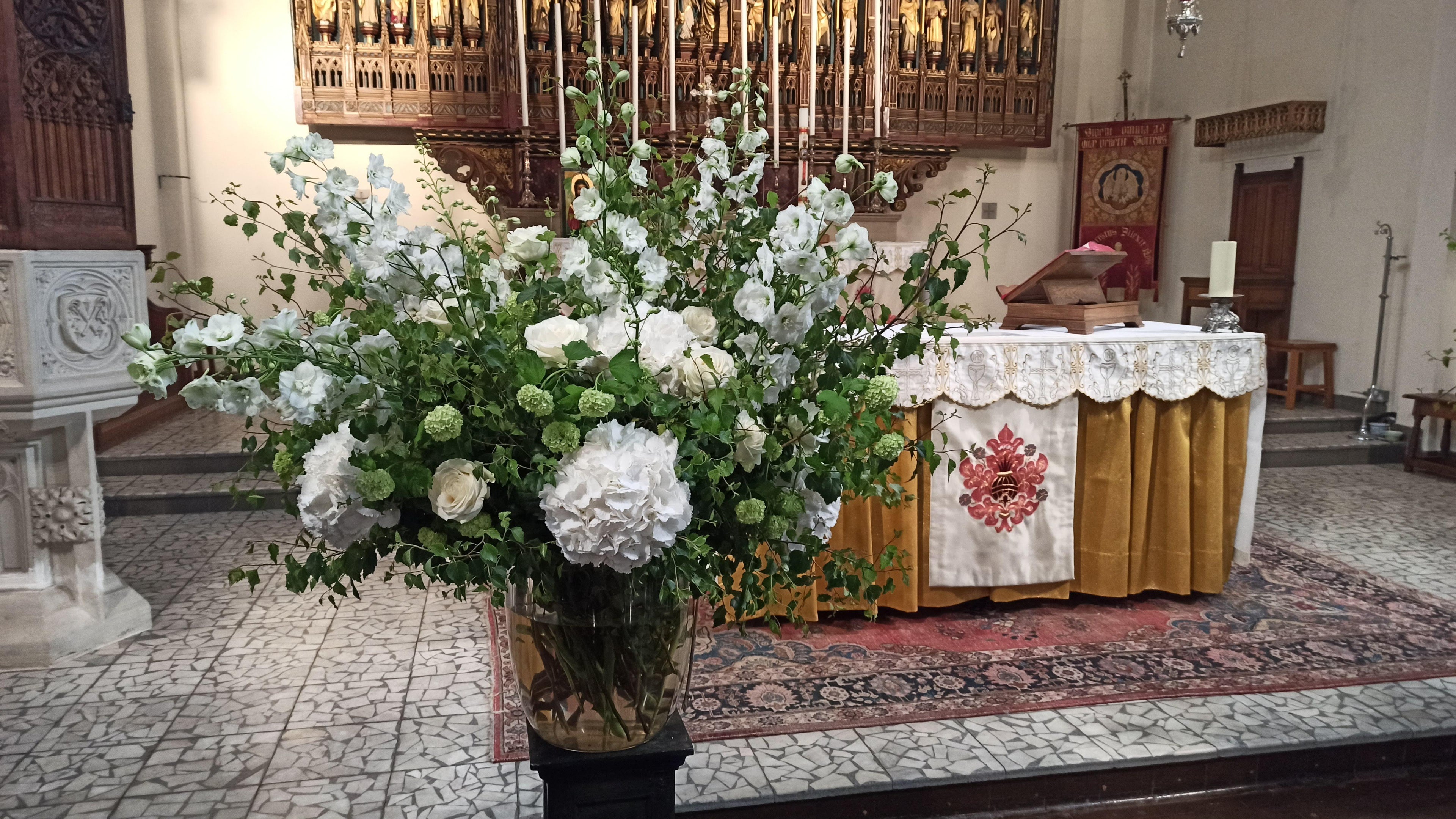 A large alter arrangemnt in a pedestal vase with white hydrangeas, white delphinium and roses