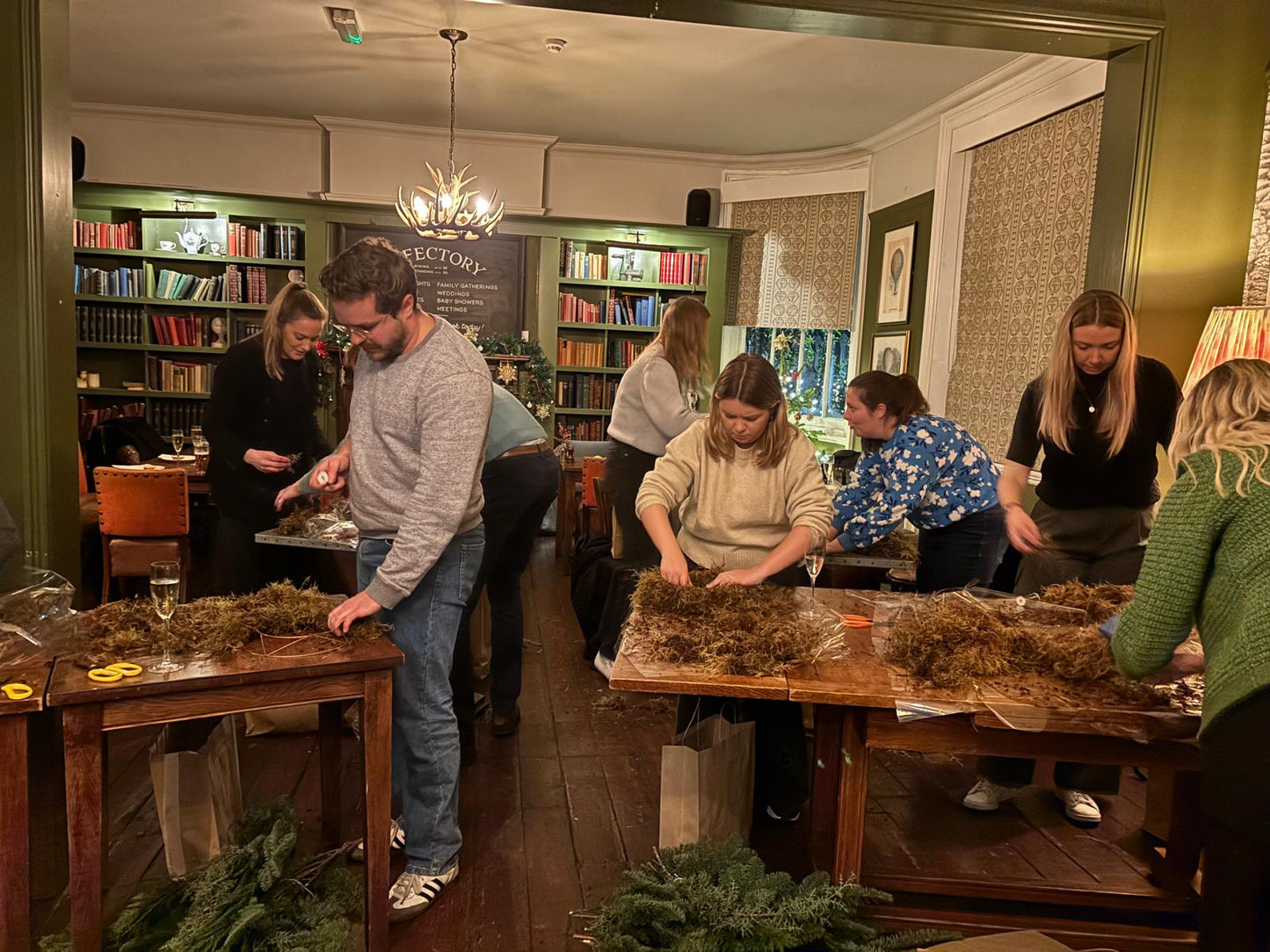 Group of people working on wreaths in a room with bookshelves and festive decorations.