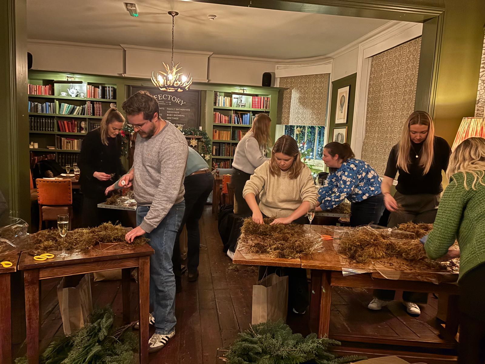 Group of people working on wreaths in a room with bookshelves and festive decorations.